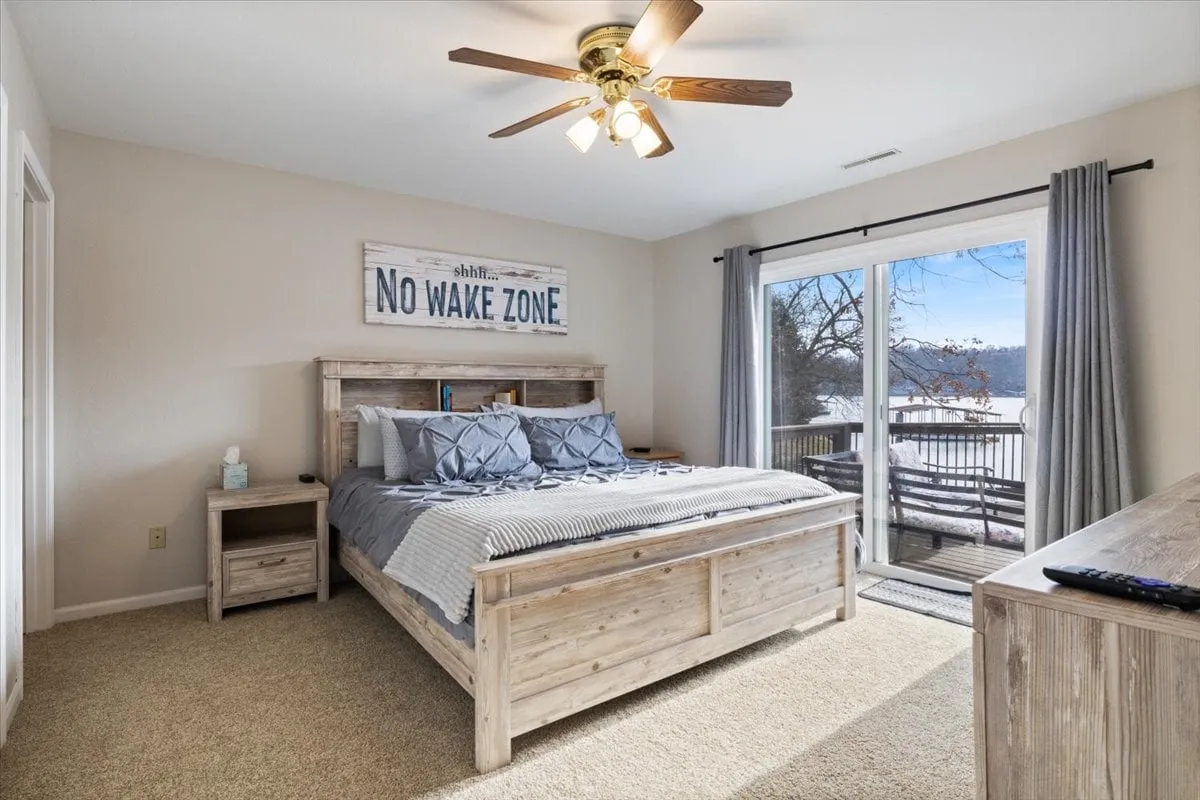 A serene bedroom featuring a plush bed with striped bedding, a wooden headboard, and large windows that provide a stunning view of the lake. The space is bathed in natural light and accented by a sign reading 'No Wake Zone,' emphasizing the tranquil, restful vibe. The room offers direct access to the outdoors, inviting lake breezes and opportunities for stargazing.