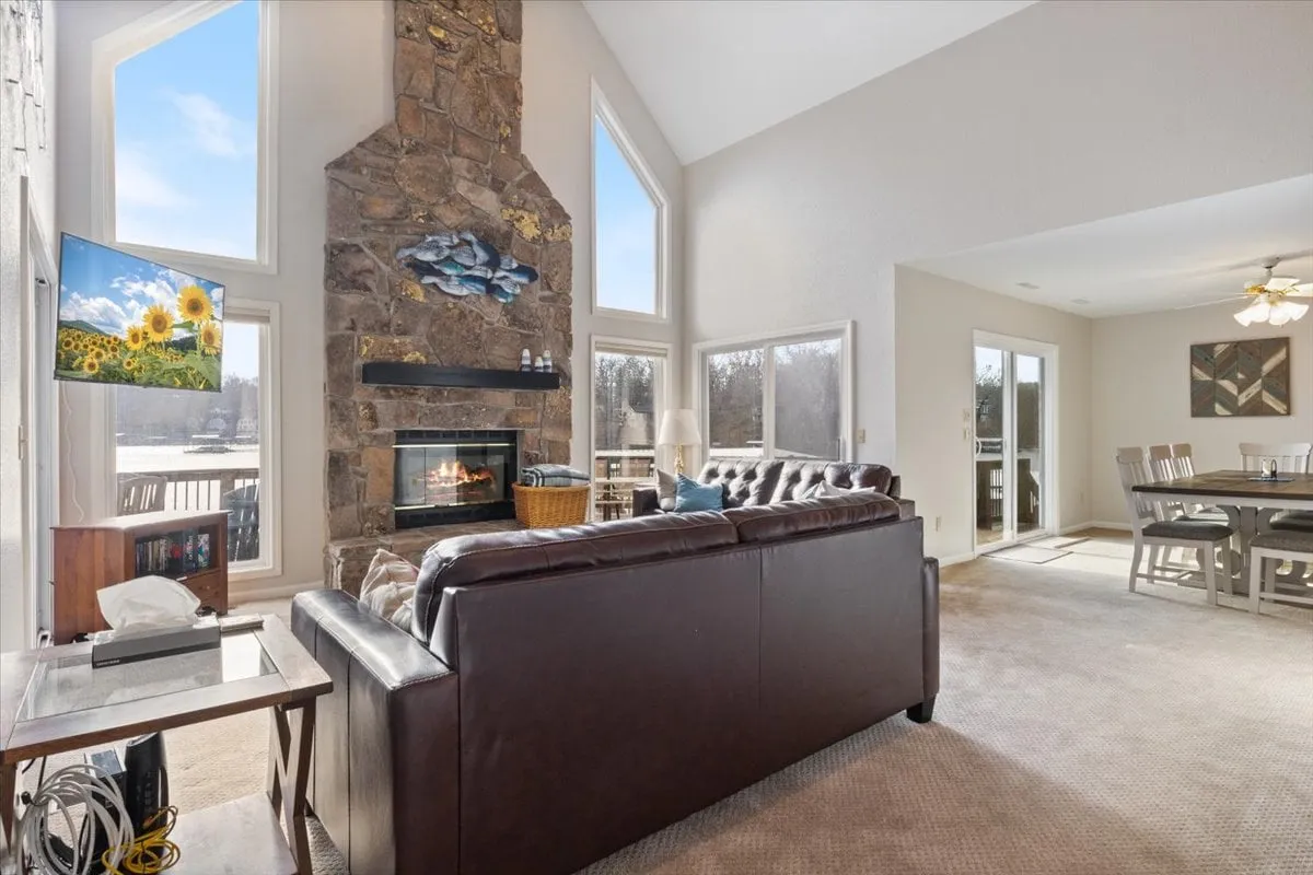 A warm, welcoming living and dining space with a stone fireplace, large windows letting in natural light, a leather couch, and an adjacent dining area. The inviting setup fosters both relaxation by the fire and communal meals, all set against the backdrop of lake scenery.