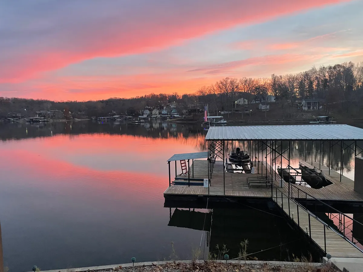 A tranquil dock scene at sunset, with water reflecting the sky’s colors, trees framing the view, and distant houses softly illuminated. The peaceful atmosphere invites relaxation and quiet moments by the water.