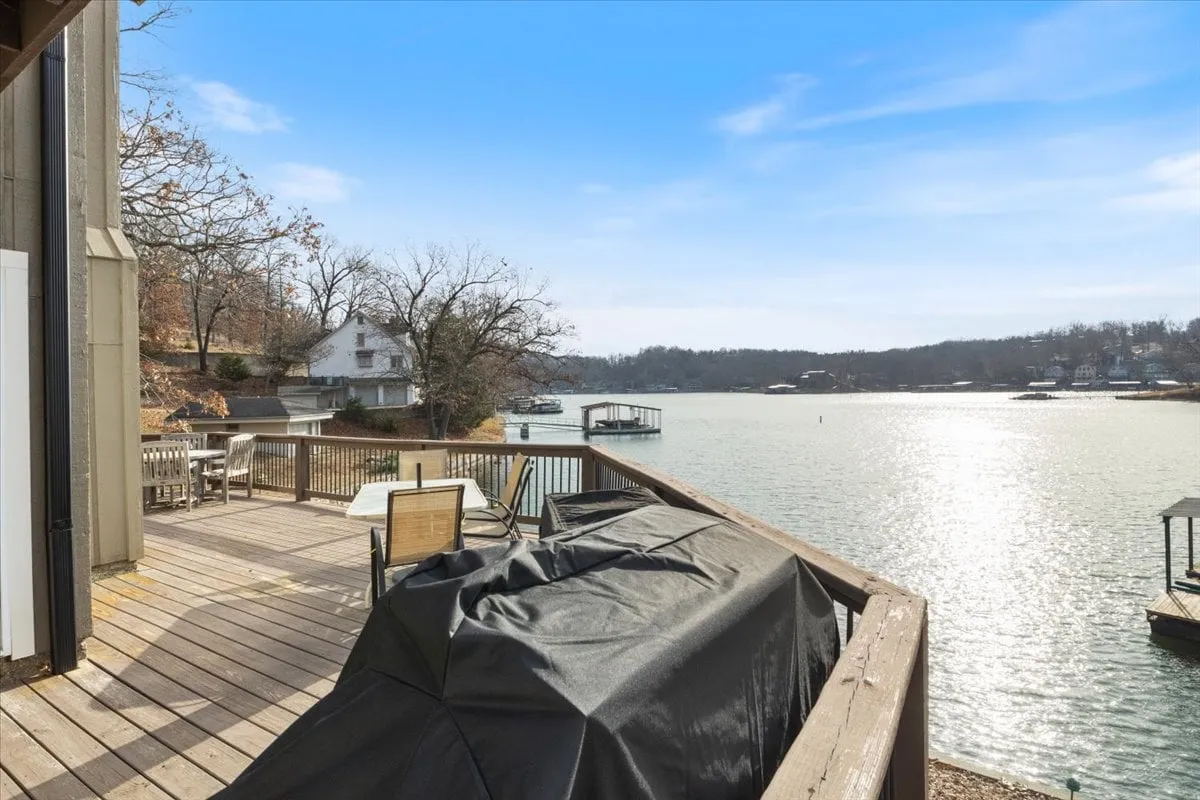 A spacious wooden deck overlooks the tranquil lake, complete with comfortable outdoor seating, a grill, and panoramic water views. The area is surrounded by lush trees, creating a serene but social outdoor setting ideal for group gatherings, morning coffee, or sunset toasts.