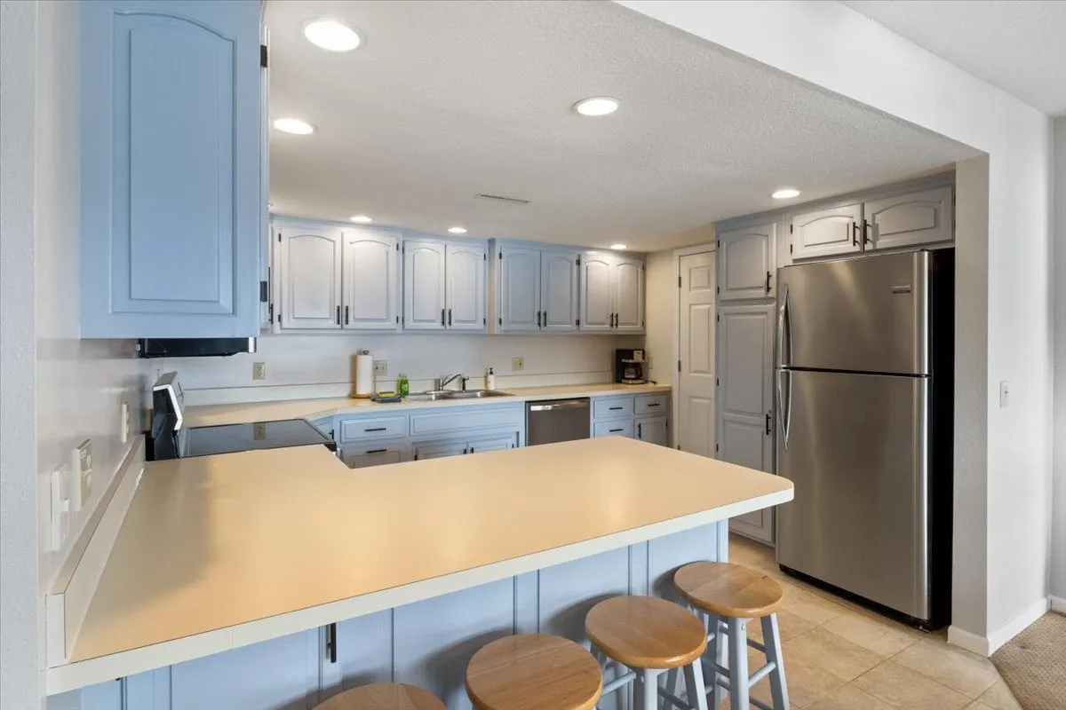 A bright, cheerful kitchen with light blue cabinetry, a central island, stainless steel refrigerator, and bar stools at the breakfast bar. Ample daylight streams in, giving the space a fresh and inviting feel, and highlighting the breakfast bar perfect for casual meals or socializing during meal prep.