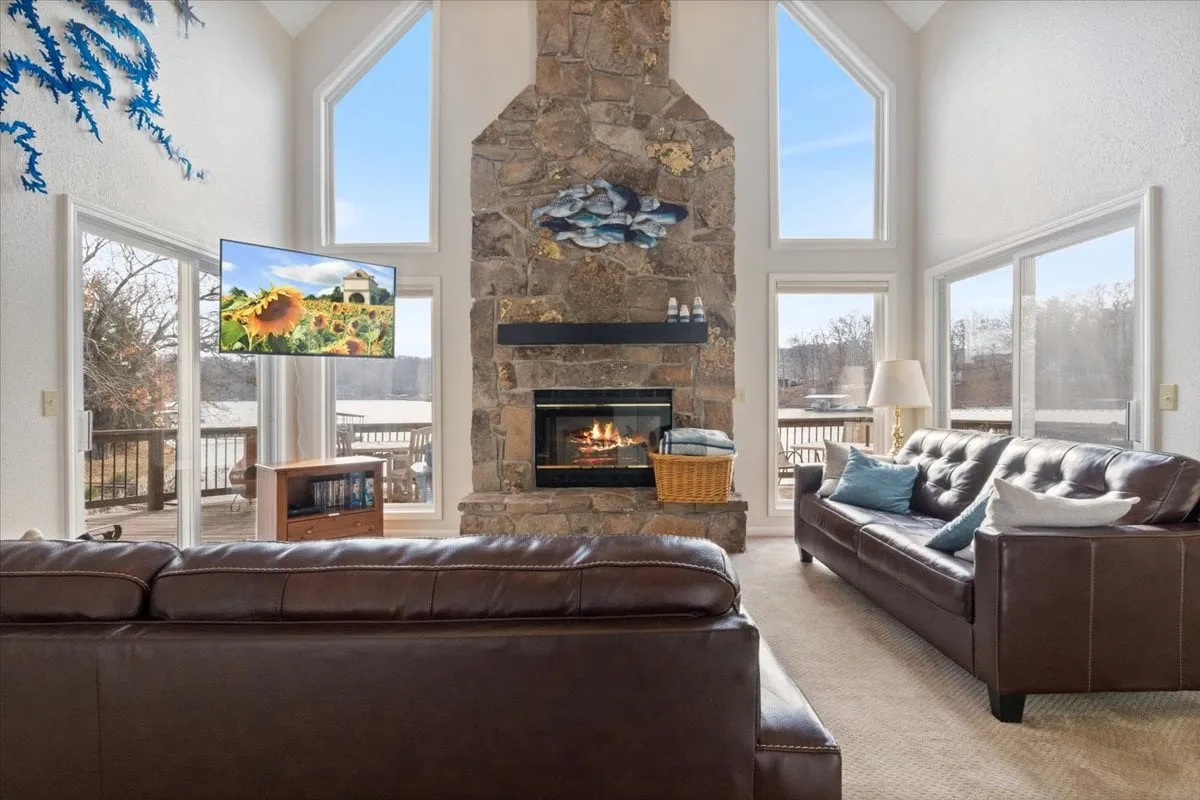 A spacious living area anchored by a large stone fireplace, with a TV displaying vibrant images, and expansive windows framing stunning views of the outdoor deck and lake. Comfortable seating invites relaxation and togetherness, and the open sightlines make the space feel bright and connected to nature.