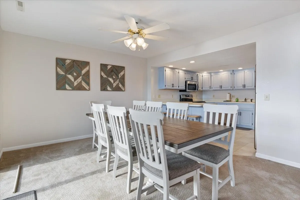A cozy dining space featuring a generous table set for a group, with a view into the adjacent kitchen. The room is decorated with homey touches such as wall art and a ceiling fan, and the atmosphere is welcoming and comfortable—ideal for social gatherings or festive dinners.