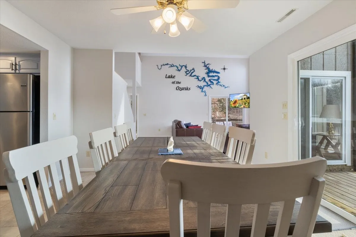 A long wooden dining table sits beneath decorative lighting, with a spacious kitchen nearby and an outdoor view visible through glass doors. The setting is ideal for family dinners, game nights, and casual conversation, with the decks just steps away.