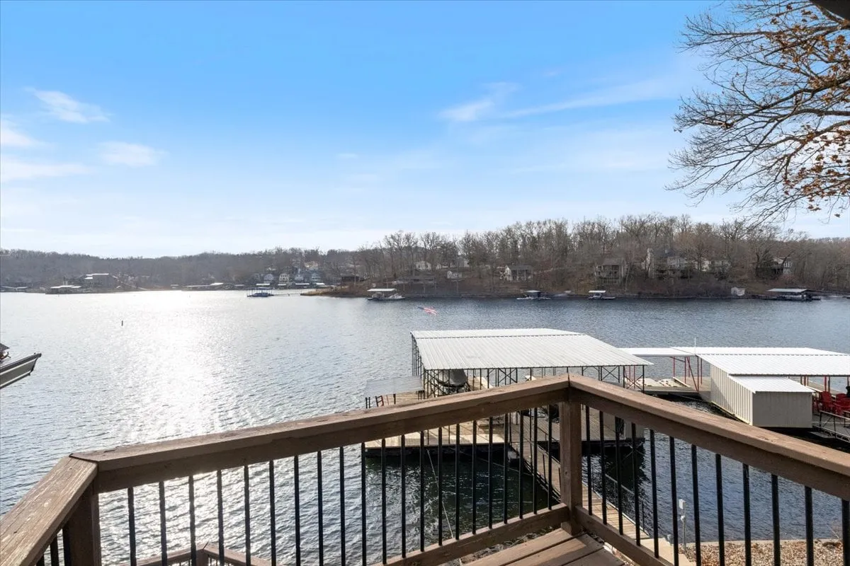 A scenic view from the dock, with calm lake waters, boats, and a wooden railing. The background includes trees and distant houses, evoking a peaceful lakeside vibe ideal for strolls, swimming, or moments of reflection by the water.