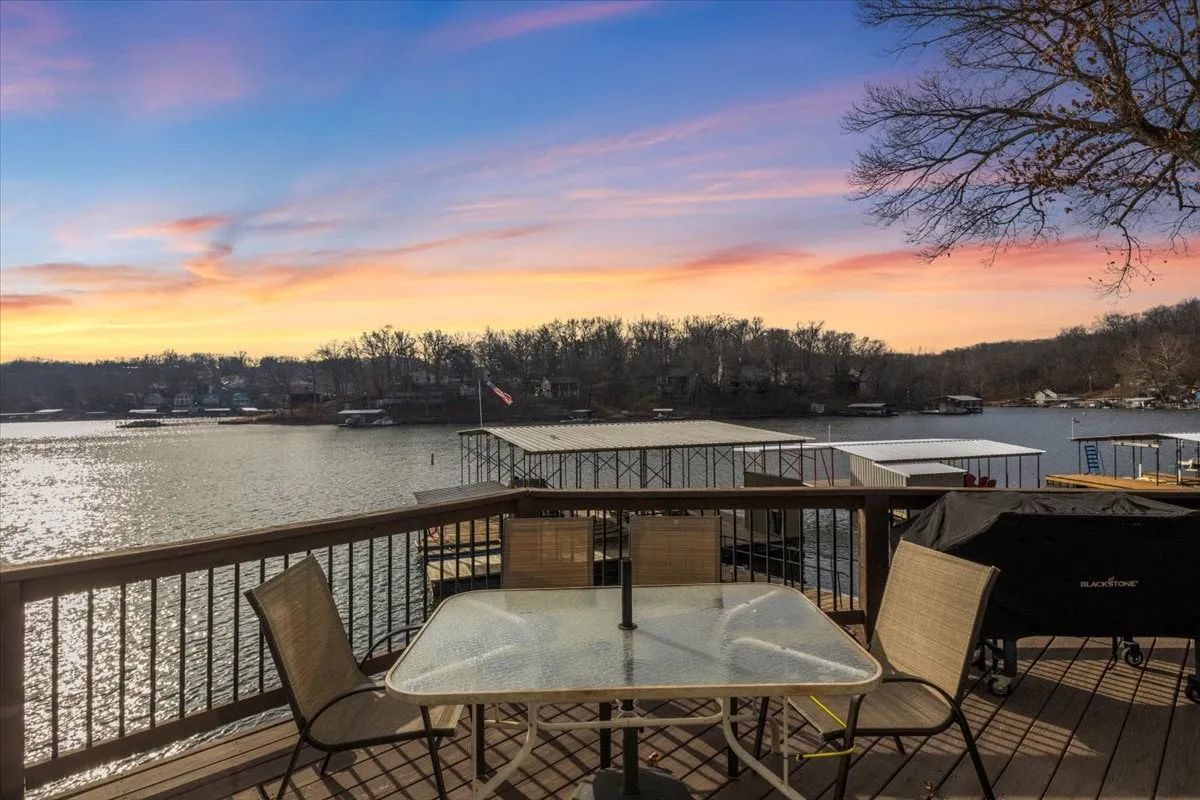 The hero image showcases an inviting outdoor deck with comfortable seating, set against sweeping lake views and a glowing sunset sky. In the distance, other lakeside homes dot the shoreline, while the tranquil water and the vibrant colors of dusk create a peaceful, welcoming ambiance.